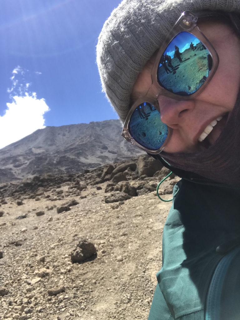 Alyson leaning into the frame excidetly with Mount Kilimanjaro in the background. She is wearing sunglasses and a winter hat.